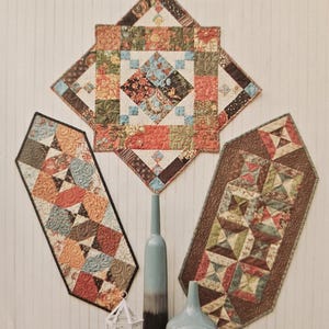 May include: Three colorful quilted table runners and a decorative centerpiece. The runners are made from 5-inch charm squares in various patterns and colors, including floral and geometric designs. The text reads "Runners designed for 5" charm squares."