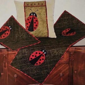May include: A decorative set featuring a ladybug theme. The set includes a pillow and several square and rectangular pieces, all with a dark green quilted background and a large red and black ladybug applique. The edges are trimmed in red.