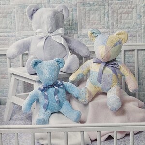 May include: Three plush teddy bears in soft pastel colors. One is light blue, one is patchwork, and the other is a pale lavender. Each bear has a satin ribbon bow around its neck. The bears are sitting on a white stool and a pink blanket.