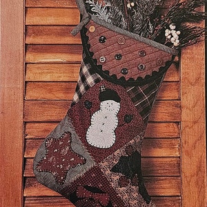 May include: A patchwork Christmas stocking with a snowman design, featuring a variety of fabrics in brown, red, and black. The stocking is decorated with buttons and is filled with a sprig of evergreen.