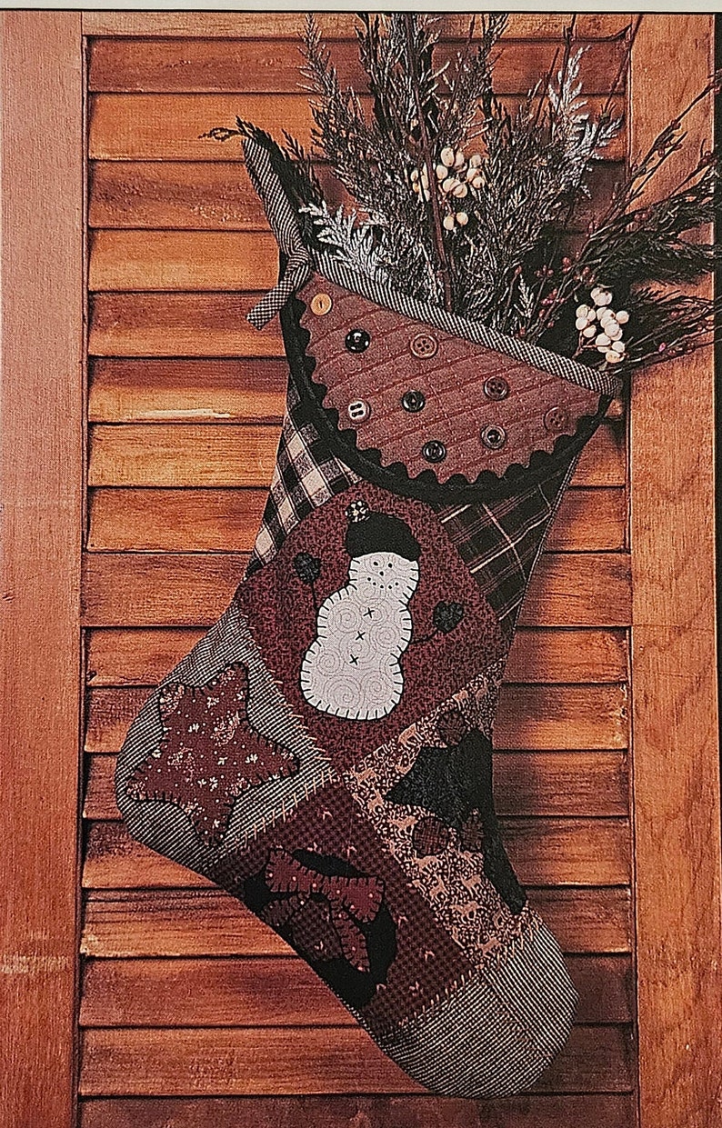 May include: A patchwork Christmas stocking with a snowman design, featuring a variety of fabrics in brown, red, and black. The stocking is decorated with buttons and is filled with a sprig of evergreen.