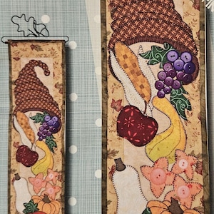 May include: Two rectangular fabric wall hangings featuring a gnome design with autumn elements. The gnomes have brown hats, and the artwork includes pumpkins, fruit, and leaves in shades of orange, yellow, and purple.