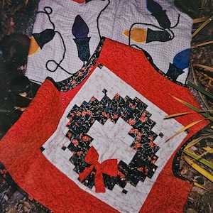 May include: A quilted Christmas vest and a square quilted mat. The vest is light gray with colorful Christmas lights appliqued on it. The mat has a red border and a wreath design with a red bow in the center.