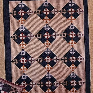 May include: A quilt with a diamond pattern in shades of blue, brown, red, and white. The quilt is made up of smaller squares that are sewn together to create the diamond pattern. The quilt is surrounded by a dark blue border.
