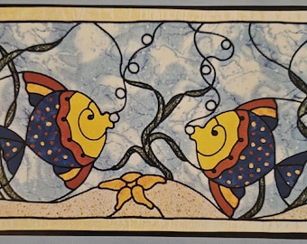 May include: Two yellow, blue, and red fish with black spots swim in a blue and white stained glass design. The fish are facing each other and are surrounded by green seaweed.