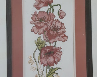 May include: A framed cross-stitch embroidery featuring three pink poppy flowers with green stems and leaves on a white background. The artwork is enclosed in a rectangular black frame with a wide, solid terracotta-colored mat border.