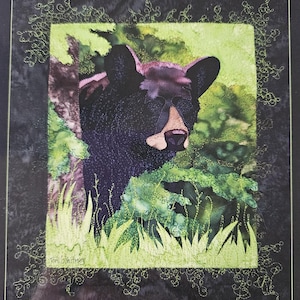 May include: A framed textile art piece featuring a black bear emerging from green foliage. The bear has a tan muzzle and is surrounded by stitched green grass and leaves. The artwork is signed by the artist, Tony Whitney.