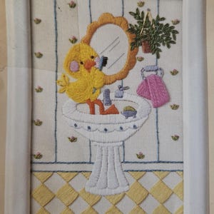 May include: A yellow embroidered chick with a pink nose is brushing its teeth in front of a mirror in a bathroom setting. The chick is standing on a white pedestal sink with a pink towel hanging on the side. There is a green plant hanging above the sink.