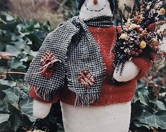 May include: A handcrafted snowman figure with a smiling face, wearing a teal knit cap, a checkered scarf, and a red sweater. The snowman holds a bouquet of dried flowers and a small decorative birdhouse. Patches adorn the scarf and body.
