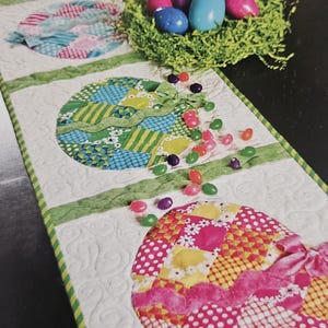 K&ouml;nnte beinhalten: Ein gesteppter Ostertischl&auml;ufer mit bunten Eierdesigns. Die Eier bestehen aus Patchwork-Stoff in verschiedenen Mustern und Farben, mit gr&uuml;ner Bandverzierung. Ein Nest mit bunten Eiern und verstreuten Geleebohnen vervollst&auml;ndigt die festliche Szene.