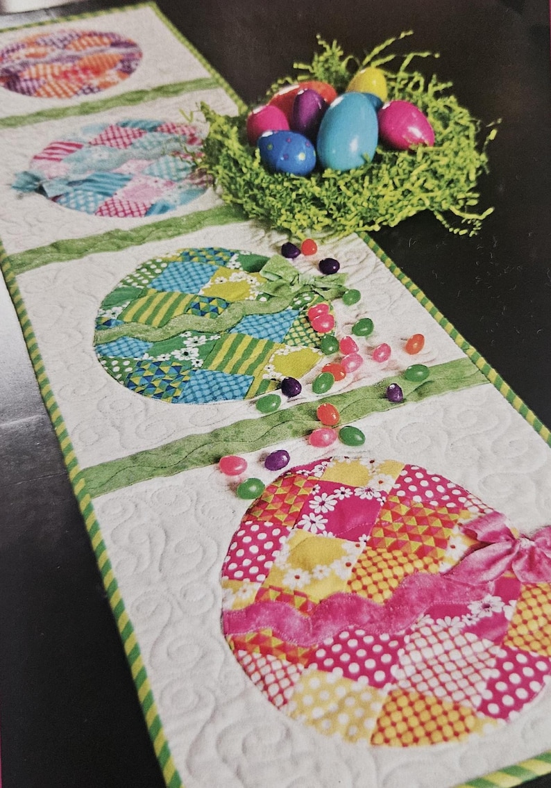 K&ouml;nnte beinhalten: Ein gesteppter Ostertischl&auml;ufer mit bunten Eierdesigns. Die Eier bestehen aus Patchwork-Stoff in verschiedenen Mustern und Farben, mit gr&uuml;ner Bandverzierung. Ein Nest mit bunten Eiern und verstreuten Geleebohnen vervollst&auml;ndigt die festliche Szene.