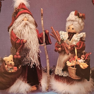 May include: A pair of handmade Christmas ornaments depicting Santa Claus and Mrs. Claus. Santa is holding a wooden staff and a bag of toys. Mrs. Claus is holding a basket of toys and a bundle of cinnamon sticks.