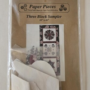 May include: A package containing paper pieces for paper piecing quilting. The package includes a brown card with the text "Paper Pieces Precut Paper Shapes for Paper Piece Quiltmaking Three Block Sampler 18" x 46"" and a picture of three quilt blocks. The package also includes white paper pieces.