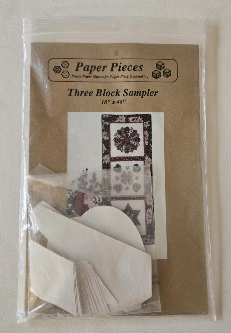 May include: A package containing paper pieces for paper piecing quilting. The package includes a brown card with the text "Paper Pieces Precut Paper Shapes for Paper Piece Quiltmaking Three Block Sampler 18" x 46"" and a picture of three quilt blocks. The package also includes white paper pieces.
