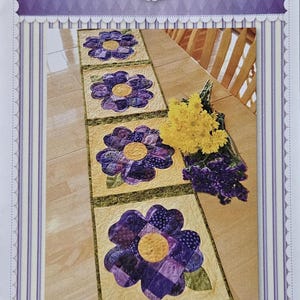 Op de afbeelding: Een patchwork tafelloper met grote paarse viooltjesbloemmotieven en gele centra op een lichtgele achtergrond. De tafelloper wordt getoond op een houten oppervlak met bloemen op de achtergrond. De tekst "Shabby Fabrics" en "Patchwork Pansy Table Runner" zijn zichtbaar.