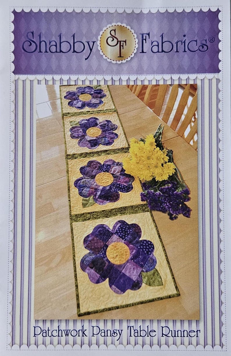 Op de afbeelding: Een patchwork tafelloper met grote paarse viooltjesbloemmotieven en gele centra op een lichtgele achtergrond. De tafelloper wordt getoond op een houten oppervlak met bloemen op de achtergrond. De tekst "Shabby Fabrics" en "Patchwork Pansy Table Runner" zijn zichtbaar.