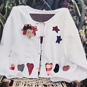 May include: White cardigan with red, blue, and white fabric hearts and stars appliqued on the front. A red-haired doll with a black bow is also appliqued on the front.