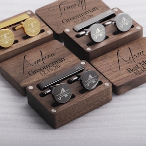 May include: Four wooden boxes with engraved names and dates, each containing a tie clip and cufflinks. The boxes are made of dark wood and have a polished finish. The cufflinks and tie clips are silver and have engraved initials.