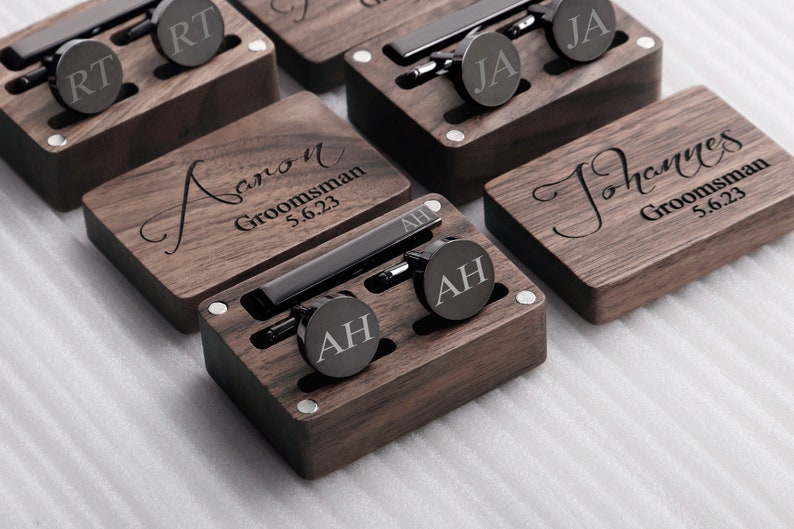 May include: Set of six wooden boxes with engraved names and dates, each containing a tie clip and cufflinks. The boxes are made of dark wood and have a polished finish.