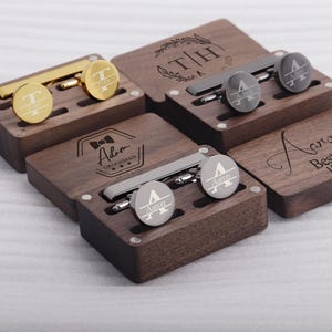 May include: Four wooden boxes with engraved initials and text. Each box contains a pair of cufflinks and a tie clip. The boxes are made of dark wood and have a polished finish.