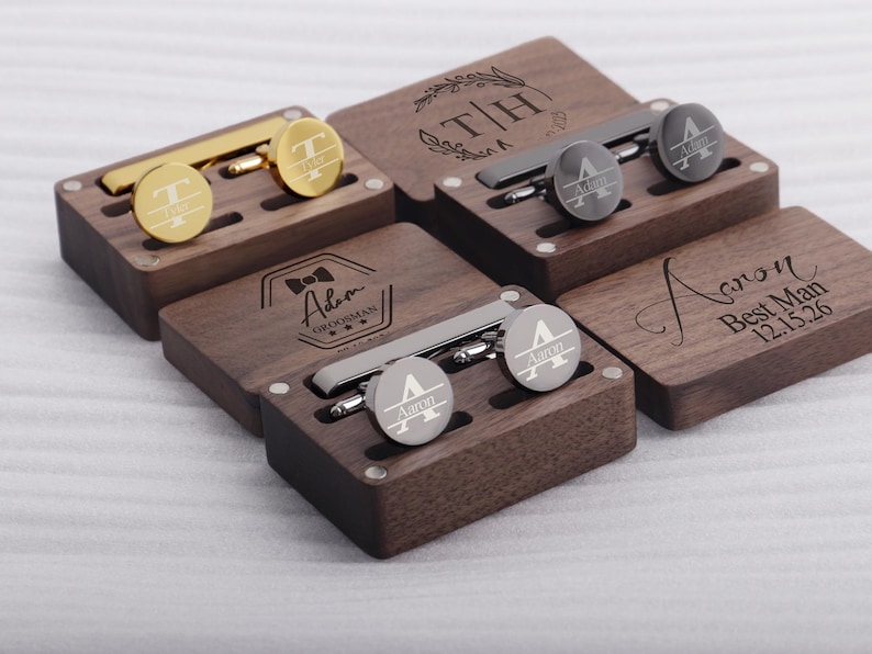 May include: Four wooden boxes with engraved initials and text. Each box contains a pair of cufflinks and a tie clip. The boxes are made of dark wood and have a polished finish.