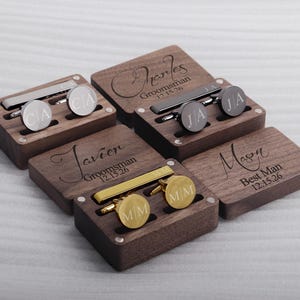 May include: Five wooden boxes with engraved names and dates. Each box contains a tie clip and cufflinks. The boxes are made of dark wood and have a polished finish.