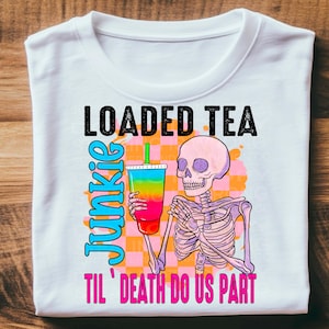 May include: White t-shirt with a graphic of a skeleton holding a colorful loaded tea with the text "Loaded Tea Junkie 'Til Death Do Us Part".
