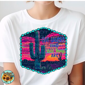 May include: A white t-shirt with a graphic design of a cactus in a desert sunset. The graphic has a turquoise border with turquoise beads and the text "Out Where The Wild Things Are".