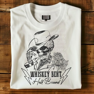 Könnte beinhalten: Weißes T-Shirt mit einem schwarz-weißen Motiv eines Skeletts mit Cowboyhut und Rose. Der Text "Whiskey Bent Hell Bound" ist darunter gedruckt. Das T-Shirt hat einen Rundhalsausschnitt und ein kleines Etikett.