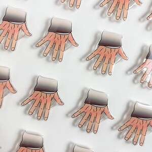 May include: A collection of acrylic pins featuring a stylized illustration of a hand with visible skin texture and veins, wearing a dark grey sleeve cuff. Each pin is identical, showing a peach-toned hand with five fingers spread apart.