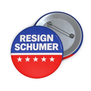 May include: A round button with the text "RESIGN SCHUMER" in white on a blue and red background. The button has a silver pin on the back. The design includes five white stars on the red portion.
