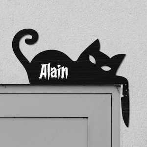 Peut inclure: Une silhouette noire de chat avec le nom "Alain" écrit dessus est montée sur un mur blanc au-dessus d'une porte grise. Un petit luminaire blanc est monté sur le mur à droite du chat.