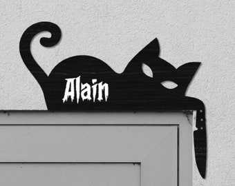 May include: A black silhouette of a cat with the name "Alain" written on it is mounted on a white wall above a gray door. A small white light fixture is mounted on the wall to the right of the cat.