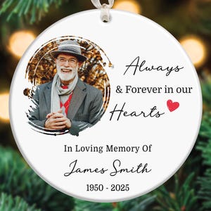 May include: A white ceramic Christmas ornament with a photo of a man in a gray jacket and hat. Text reads "Always & Forever in our Hearts" and "In Loving Memory Of James Smith 1950-2025". A red heart is next to the word "Hearts".