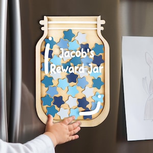May include: Wooden reward jar magnet with the text "Jacob's Reward Jar" and blue star tokens. The jar is attached to a stainless steel refrigerator. A child's hand reaches towards the jar.