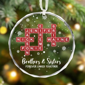 May include: A clear, round Christmas bauble with a white ribbon. It displays names in red tiles, like a crossword, with the text "Brothers & Sisters" and "FOREVER LINKED TOGETHER". Snowflakes are scattered across the surface of the bauble.