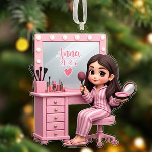 May include: Pink Christmas ornament featuring a cartoon girl in striped pajamas at a vanity. The vanity has a mirror with the name "Anna" and the year "2025". A heart is below the year. The ornament is hanging from a white ribbon.
