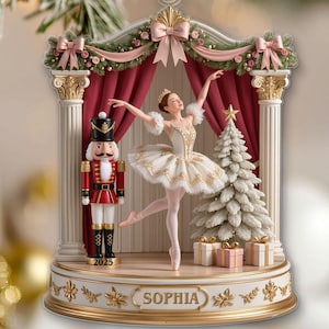 Könnte beinhalten: Weihnachtsschmuck mit einer Ballerina auf einer Bühne mit einem Nussknacker und einem Weihnachtsbaum. Der Schmuck hat eine weiße Basis mit dem Namen "SOPHIA" und dem Jahr "2025". Die Ballerina trägt ein weißes Tutu und eine Krone. Die Bühne hat rote Vorhänge und weiße Säulen.