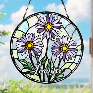 Może przedstawiać: Okrągła witrażowa zawieszka do okna z trzema fioletowymi stokrotkami, żółtymi środkami i zielonymi liśćmi. Imię "Jennifer" jest napisane na dole. Okno jest oprawione w czarną ramę i wisi na łańcuchu.