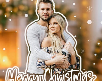 May include: A clear acrylic Christmas ornament featuring a couple embracing, with the words "Merry Christmas" in a cursive font. The ornament is suspended by a white ribbon. The background includes a Christmas tree and festive lights.