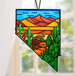 May include: A stained glass ornament in the shape of Nevada, featuring a desert landscape. The design includes red mountains, cacti, and lizards, with a blue sky and a black chain for hanging. The ornament is a vibrant piece of art.