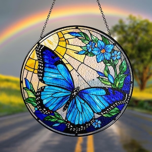 May include: A stained glass suncatcher featuring a blue butterfly with black markings and a floral design with blue flowers. The suncatcher is round and has a black metal frame.