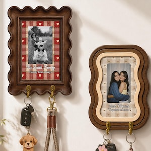Op de afbeelding: Twee rechthoekige fotolijsten voor aan de muur met golvende randen en geïntegreerde messingkleurige haken. De linkerlijst heeft een rood geruite achtergrond met de tekst "The Smith Family" en "Family first, always." De rechterlijst heeft een beige geruite achtergrond met de tekst "Best Friends" en "Together is better." Beide lijsten houden sleutels vast.