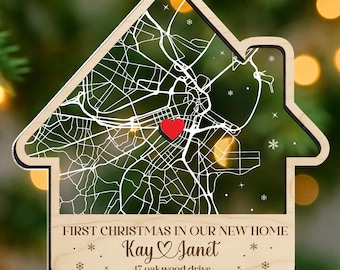 May include: Wooden house-shaped Christmas ornament with a map design and a red heart. The ornament has the text "FIRST CHRISTMAS IN OUR NEW HOME" and "Kay & Janet, 47 oakwood drive." It is decorated with a red and white checkered bow.
