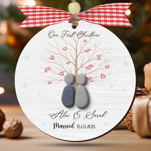 May include: A round wooden Christmas ornament with the text "Our First Christmas". The ornament features a tree design with red berries and two pebble figures. The names "Alex & Sarah" and the date "15.09.2025" are also included. A red and white ribbon is attached.
