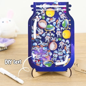 May include: A blue and white jar with a space theme. The jar is filled with astronaut and space-themed stickers. The jar is on a wooden stand. The text "Thank you for being part of my story" is on a wooden tag.