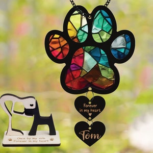 May include: A stained glass paw print suncatcher with a rainbow of colors. It hangs from a black chain and has two black heart-shaped charms with the text "Forever in my heart" and "Tom".