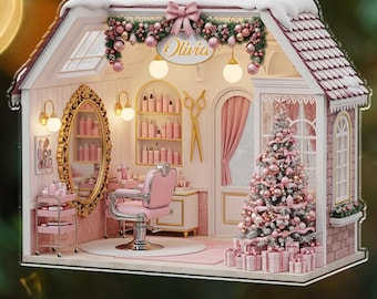 May include: A festive Christmas tree ornament in the shape of a salon. The interior is decorated in pink and white, with a chair, mirror, and Christmas tree. The name "Olivia" is displayed. A red and white gingham bow adorns the top of the ornament.