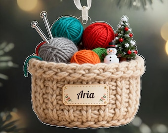 May include: A beige knitted basket Christmas ornament filled with colorful yarn balls, knitting needles, a miniature Christmas tree, and a snowman. The ornament has a tag that reads "Aria".