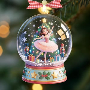 May include: A festive Christmas snow globe ornament. Inside, a ballerina in a pink tutu dances before a decorated Christmas tree. The base is adorned with stars and holly. The name "Amelia" is displayed on the base of the ornament.